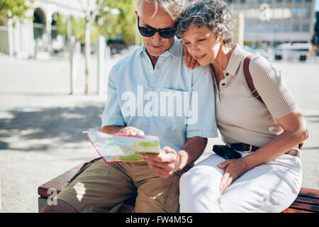 Coppia senior in cerca di destinazione su una mappa della città. Due anziani attiva la lettura di una mappa insieme mentre seduti all'aperto su un banco di lavoro Foto Stock