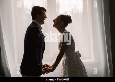 Happy sposi novelli ridere indoor. Bruna sposa e lo sposo supporto vicino alla finestra sagome di felice Foto Stock