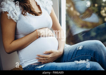 Primo piano del ventre di una futura madre. Incinta bruna ragazza seduta sul davanzale, tenendo il suo stomaco al di fuori, i verdi, i Foto Stock