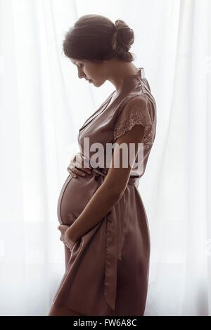 Silhouette di donna incinta in una rosa negligee in piedi contro la finestra. Vista laterale di felice donna incinta guardando pancia Foto Stock