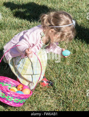 Una bambina caucasica di due anni cova uova di Pasqua, raccogliendone una dall'erba. Foto Stock