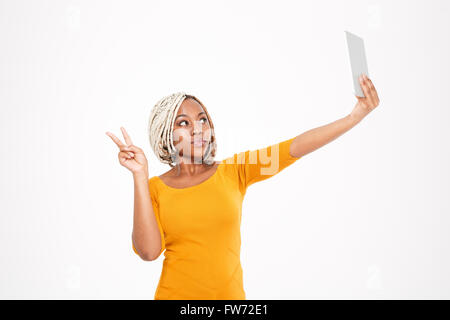 Giocoso carino giovane africano donna americana rendendo selfie con tablet e mostrando segno di vittoria su sfondo bianco Foto Stock