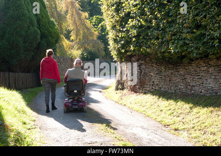 Vista posteriore di un uomo anziano con una mobilità scooter con il suo accompagnatore a camminare accanto a lui Foto Stock