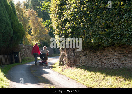 Vista posteriore di un uomo anziano con una mobilità scooter con il suo accompagnatore a camminare accanto a lui Foto Stock