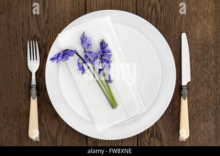 Tavola di Cena luogo con impostazione del colore bianco porcellana piatti, bluebell fiori, antichi posate e tovagliolo di lino su sfondo di quercia. Foto Stock