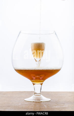 La birra è di versare in un bicchiere che è in un altro enorme di vetro, la creazione di schiuma e bolle sulla texture in legno nella parte inferiore di un Foto Stock