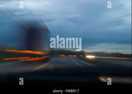 La vista attraverso il finestrino di un'automobile come si impadronisce di un camion o carrello a velocità che mostra chiaramente con l'aiuto di una lenta velocità di otturatore sulla fotocamera fotografi le striature del camion e le vetture davanti quando scende la notte su questa strada bbusy. Foto Stock