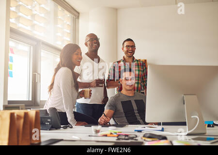 Casualmente vestito ridendo donna e tre uomini che tengono le bevande attorno al computer con stilo e compressa sulla scrivania in ufficio Foto Stock