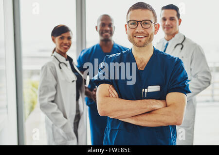 Medico di fiducia nella parte anteriore del gruppo sorridente alla fotocamera, indossando abiti da chirurgo Foto Stock