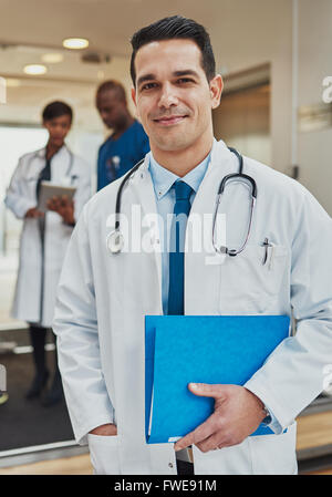 Gentile fiducioso ispanico medico con un file del paziente nelle sue mani indossando un camice e stetoscopio guardando la telecamera wit Foto Stock