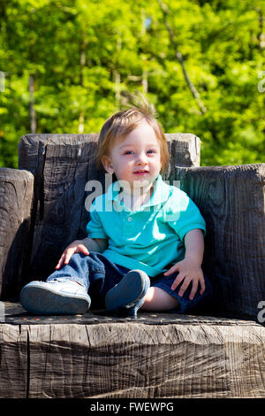 Un simpatico ragazzo si siede su un moncone intagliato in una sede in un parco all'aperto per questo grazioso e semplice ritratto. Foto Stock