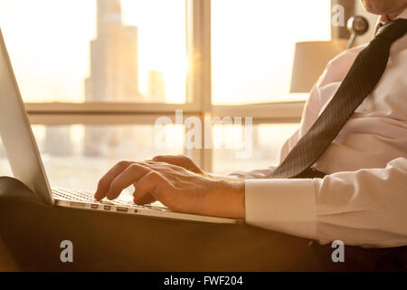 Bracci del bel giovane imprenditore formale che indossa una camicia bianca e cravatta digitando su laptop seduta sul letto in camera moderna Foto Stock