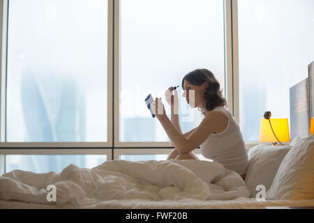 Ritratto di giovane donna seduta sul letto disfatto dopo il risveglio e mettere il mascara. Caucasian modello femminile Foto Stock