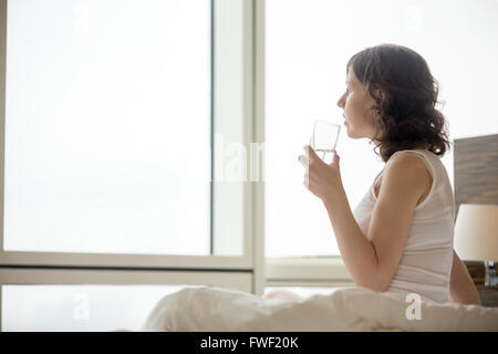 Stile di vita Ritratto di giovane donna seduta sul letto di acqua potabile dopo alzarmi. Caucasian modello femminile rilassante al mattino Foto Stock