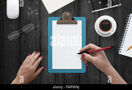 Femmina di scrittura a mano su notebook circondato con accessori per ufficio. Vista superiore della scrivania. Caffè, bicchieri, matita, mouse, carta Foto Stock