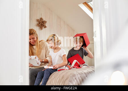 La mattina di Natale in una casa famiglia. Una madre e due bambini seduti su un letto apertura presenta. Foto Stock