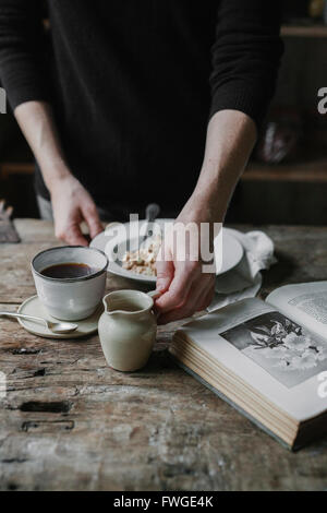 Una persona in una tabella con una tazza di caffè, ciotola di muesli e un libro aperto. Foto Stock