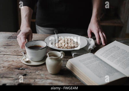 Una persona in una tabella con una tazza di caffè, ciotola di muesli e un libro aperto. Foto Stock