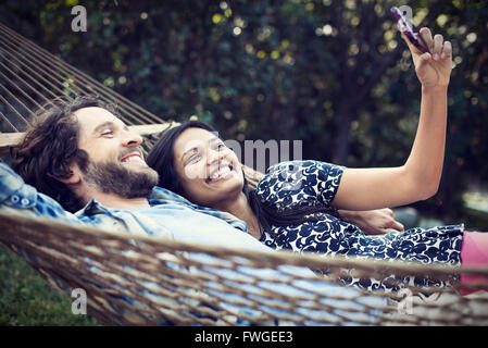 Un giovane, un giovane uomo e donna giaceva in una grande amaca in giardino, prendendo un selfy degli stessi. Foto Stock