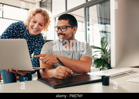 Due giovani graphic designer che lavorano insieme con il collega di sesso femminile che mostra qualcosa di uomo seduto al suo des. Le persone creative di co Foto Stock