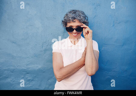 Ritratto di sorridere donna matura peeking over Sunglasses contro sfondo blu. Bel mezzo le donne più anziane guardando la fotocamera Foto Stock