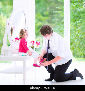 Giovane padre felice in un business suit e barra di aiutare la sua figlia piccola, carina curly toddler ragazza a mettere su un pattino Foto Stock