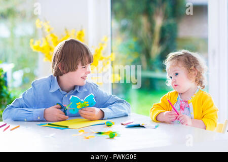 Due bambini felici, scuola ragazzo di età e poco toddler girl divertirsi insieme pittura e taglio a farfalla di carta prima di pasqua Foto Stock