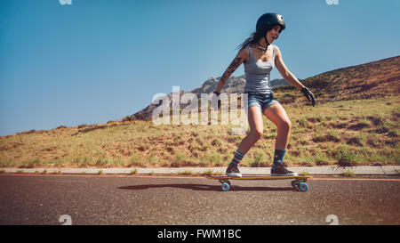 Giovane donna lo skateboard con una strada in discesa. Guidatore di skateboard femmina indossando il casco il pattinaggio su strada rurale. Foto Stock