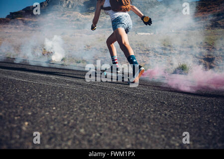 Immagine ritagliata della giovane donna lo skateboard su una strada. Skateboard con fumo colorato granata. Foto Stock