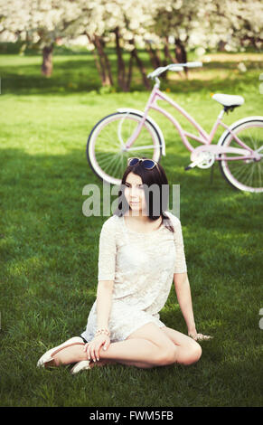 Attraente ragazza giovane con la bicicletta rosa trascorrere il tempo libero nel parco verde a bright primavera o estate giorno. Modello indossa un sundress e occhiali da sole, il suo umore è grande e lei è bellissima Foto Stock