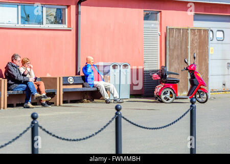 Simrishamn, Svezia - Aprile 1, 2016: Senior uomo e giovane sit crogiolarsi al sole questa calda giornata di primavera. Parcheggiate wheelchai elettrico Foto Stock