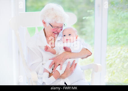Nonna amorevole di cantare una canzone al suo neonato nipote seduto in un bianco sedia a dondolo accanto ad un grande giardino vista finestra Foto Stock