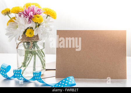 La molla bouquet di fiori in un vaso vaso con carta kraft nota, Busta e puntini blu nastro su un legno bianco sfondo rustico Foto Stock