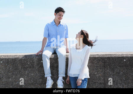 Un giovane giapponese giovane seduto sul muro di cemento in riva al mare Foto Stock