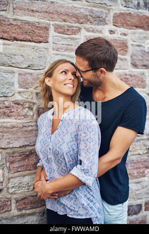Mezzo Corpo colpo di un uomo che abbraccia la sua donna da dietro romanticamente e sorridente ogni altra contro un muro di mattoni dello sfondo. Foto Stock