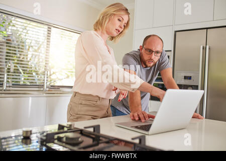 Ritratto di Coppia matura lavorando insieme su un computer portatile in cucina. Matura in piedi dal banco di cucina, con la donna pointi Foto Stock