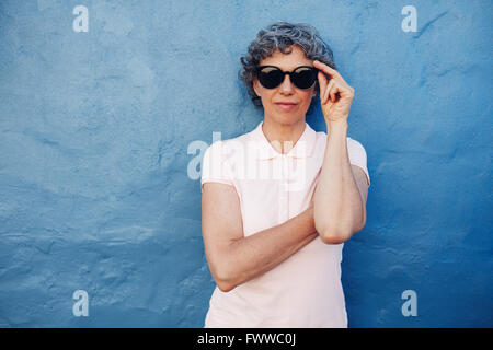 Ritratto di eleganti donna matura con occhiali da sole contro lo sfondo blu. Femmina caucasica in piedi contro la parete di blu. Foto Stock