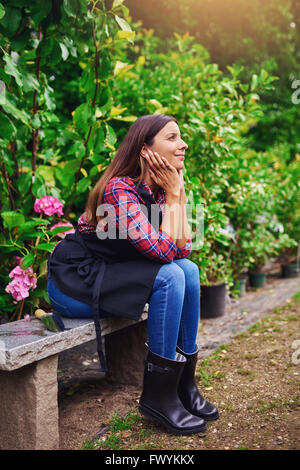 Piuttosto giovane lavoratore vivaio seduto su una panca in legno fantasticando con un sorriso di piacere sul suo viso come ella si prende una pausa in t Foto Stock