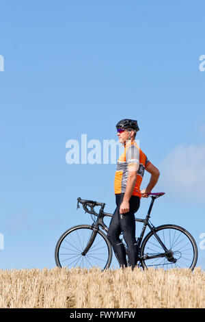 Uomo in camicia arancione in giallo raccolte campo di mais tenendo la bici in una giornata soleggiata con un cielo azzurro Foto Stock