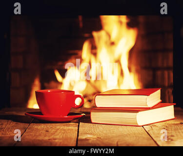 Red tazza di tè o caffè e libri antichi vicino al camino sul tavolo di legno. Inverno e vacanze di Natale concetto. Foto con retro fi Foto Stock