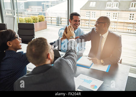 Successo aziendale multirazziale team seduti a un tavolo in un ufficio urbano il tifo e congratulandomi con ogni altro dopo un outsta Foto Stock