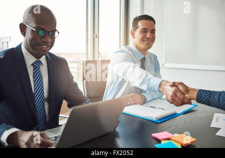 Sorridente giovane azienda ispanica l uomo e la donna si stringono la mano su un tavolo in ufficio guardato da un sorridente americano africano m Foto Stock