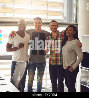 Il gruppo di quattro diversi amici vestito in abbigliamento casual e tenendo le bevande mentre sta in piedi vicino a ogni altro in ufficio con la Foto Stock