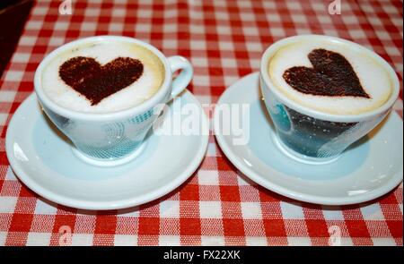 Due tazze da caffè con cuori impresse nel cappuccino con la schiuma su un rosso Gingham cafe tabella. Foto Stock