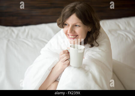 Ritratto di Allegro giovane donna seduta sul letto disfatto dopo il risveglio e di bere caffè. Modello femminile con sognante Foto Stock