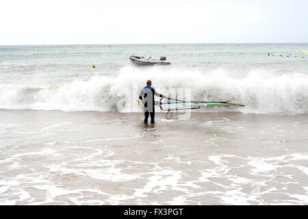 Un uomo che porta la sua tavola da surf in acqua per praticare windsurf, wind surf presso la spiaggia di Costa Teguise Lanzarote, Spagna, onde schiumose concept Foto Stock