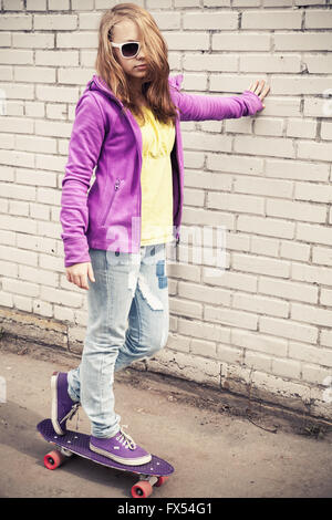 La bionda ragazza adolescente in occhiali da sole e colorato abbigliamento sportivo su uno skateboard vicino al bianco urban un muro di mattoni. La correzione delle tonalità foto Foto Stock