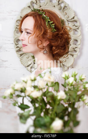 I giovani i capelli rossi sposa in una ghirlanda di verde dolcemente sorridente. Rose bianche, amore, tenerezza. Foto Stock