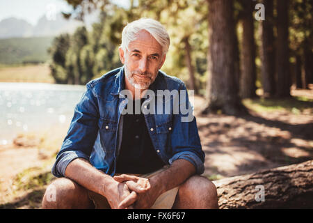 Ritratto di uomo maturo seduto vicino a un lago con lo sguardo alla telecamera. Senior uomo caucasico rilassante su un log dal lago in un giorno di estate Foto Stock