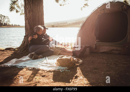 Ritratto di amorevole coppia senior campeggio nei pressi di un lago in una bella giornata d'estate. Essi sono qui seduti sotto un albero al di fuori della tenda, Foto Stock